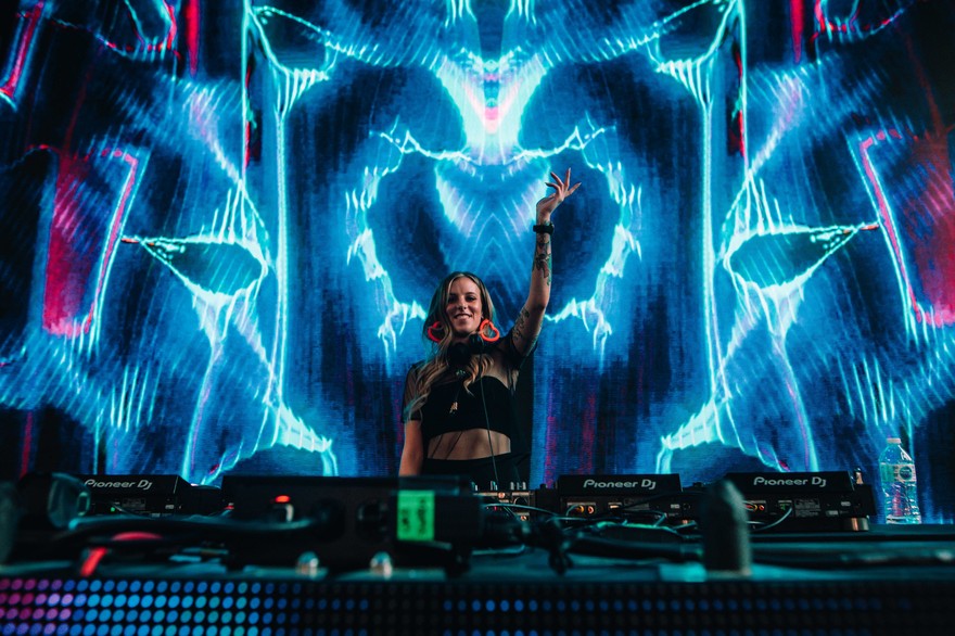 A vibrant colour photo shows Carissa Gem DJing in front of turntable with their left hand in the air and blue and red lightning-type projections behind them. 