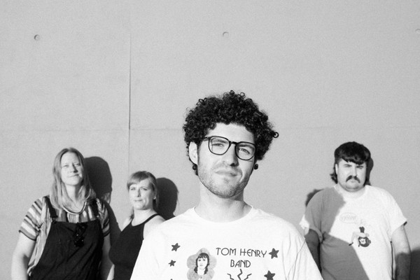 A black and white photo shows four-piece band Buddie standing in front of a white concrete wall. 