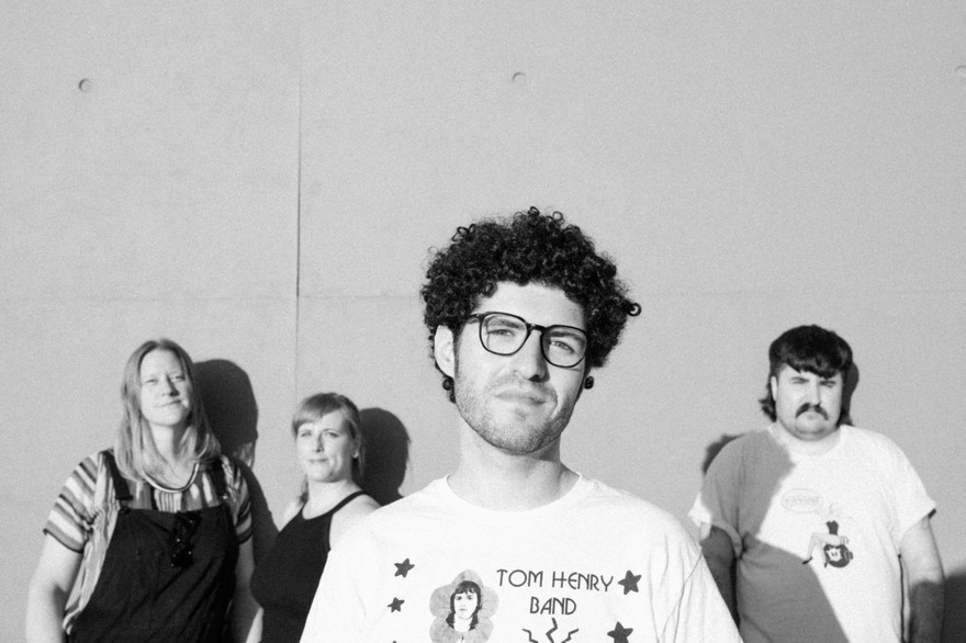 A black and white photo shows four-piece band Buddie standing in front of a white concrete wall. 