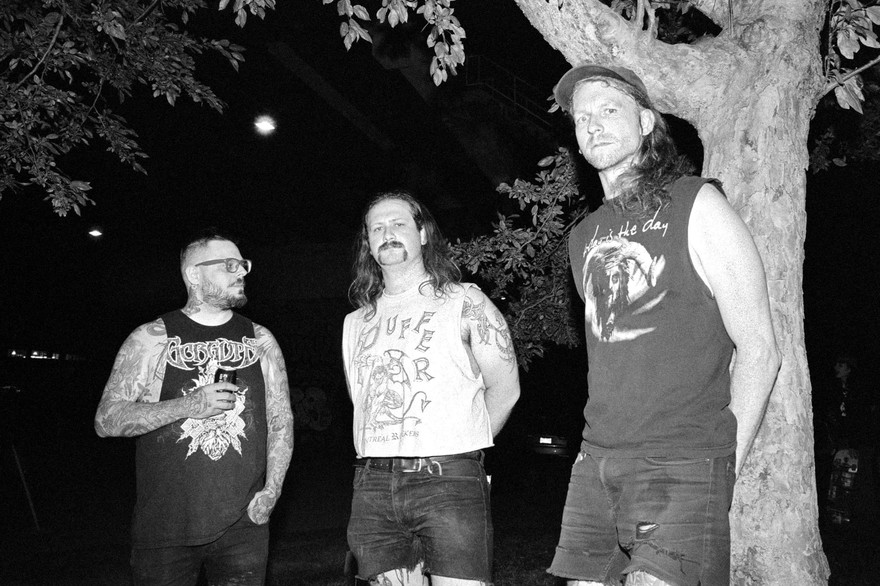A black and white photo shows three-piece group Cell Press standing outside with a large tree to the far right. 