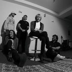 A black and white fisheye photo taken from below shows five-piece band posing inside a white room with a large patterned rug in the in foreground.