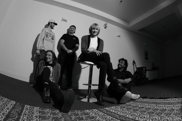 A black and white fisheye photo taken from below shows five-piece band posing inside a white room with a large patterned rug in the in foreground.
