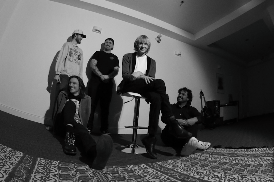 A black and white fisheye photo taken from below shows five-piece band posing inside a white room with a large patterned rug in the in foreground.