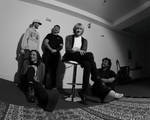 A black and white fisheye photo taken from below shows five-piece band posing inside a white room with a large patterned rug in the in foreground.