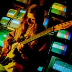 A vibrant colour photo shows artist Chic Chameleon playing guitar with long brown hair standing in front of a wall of colour TVs. 