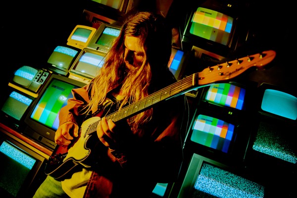 A vibrant colour photo shows artist Chic Chameleon playing guitar with long brown hair standing in front of a wall of colour TVs. 