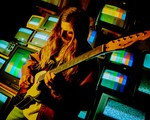 A vibrant colour photo shows artist Chic Chameleon playing guitar with long brown hair standing in front of a wall of colour TVs. 
