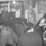 A grainy black and white photo shows four members of the group Chlorine Time Machine performing in an intimate indoor venue. 