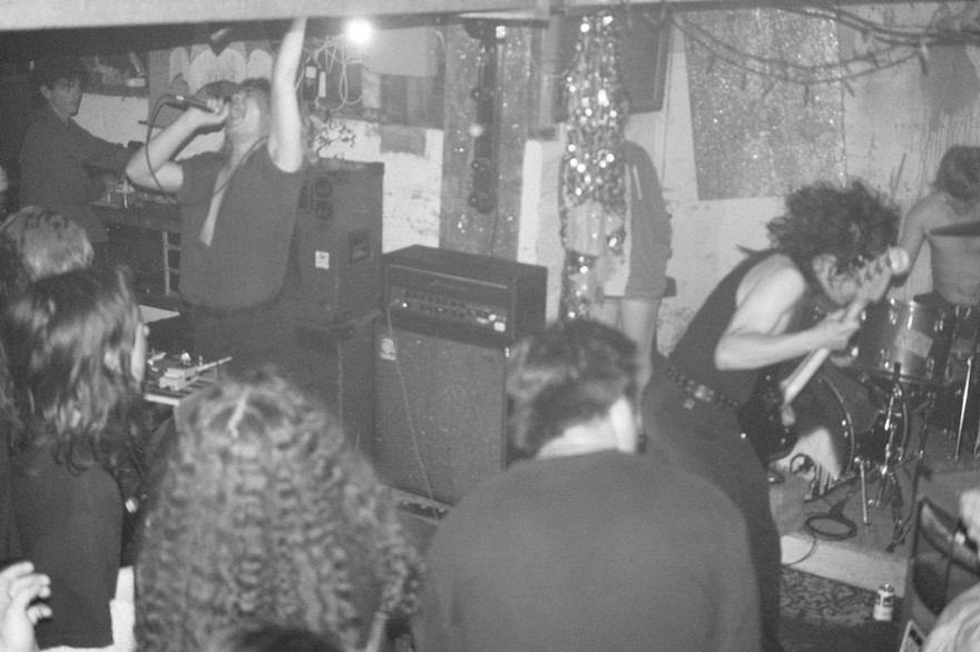 A grainy black and white photo shows four members of the group Chlorine Time Machine performing in an intimate indoor venue. 