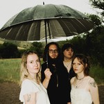 A colour photo shows four-piece group Circling Girl collectively standing under an umbrella while standing in a grassy field. 
