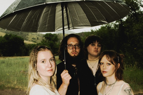 A colour photo shows four-piece group Circling Girl collectively standing under an umbrella while standing in a grassy field. 