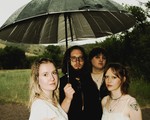 A colour photo shows four-piece group Circling Girl collectively standing under an umbrella while standing in a grassy field. 