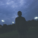 A dark grainy photo shows solo artist from Clothelines from Hell standing in a green field at dusk. Dark blue clouds fill the sky and large stadium lights are on in the background. 