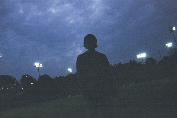 A dark grainy photo shows solo artist from Clothelines from Hell standing in a green field at dusk. Dark blue clouds fill the sky and large stadium lights are on in the background. 