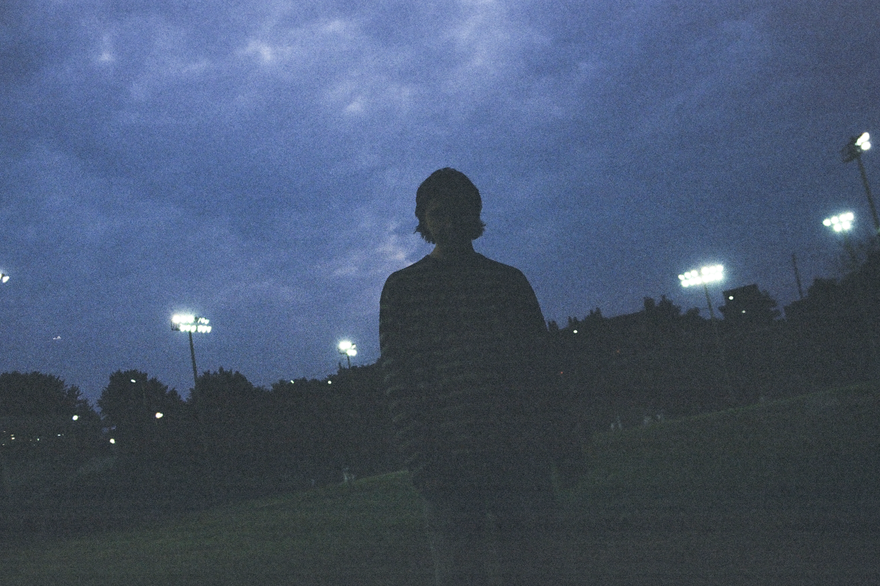 A dark grainy photo shows solo artist from Clothelines from Hell standing in a green field at dusk. Dark blue clouds fill the sky and large stadium lights are on in the background. 
