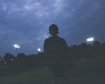 A dark grainy photo shows solo artist from Clothelines from Hell standing in a green field at dusk. Dark blue clouds fill the sky and large stadium lights are on in the background. 