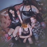 Four-piece group dead ex girlfriends are shown in a messy living room sitting on the couch and on the floor looking distraught, with a few members smoking cigarettes.
