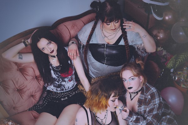 Four-piece group dead ex girlfriends are shown in a messy living room sitting on the couch and on the floor looking distraught, with a few members smoking cigarettes.