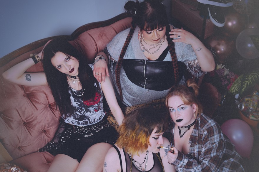 Four-piece group dead ex girlfriends are shown in a messy living room sitting on the couch and on the floor looking distraught, with a few members smoking cigarettes.