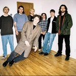 A colour photo shows seven-piece band Concrete Vehicles standing in a bedroom with hardwood floors and white walls. The member in the front is edited to look as if they are standing on a bizarre tilt as if they're falling.