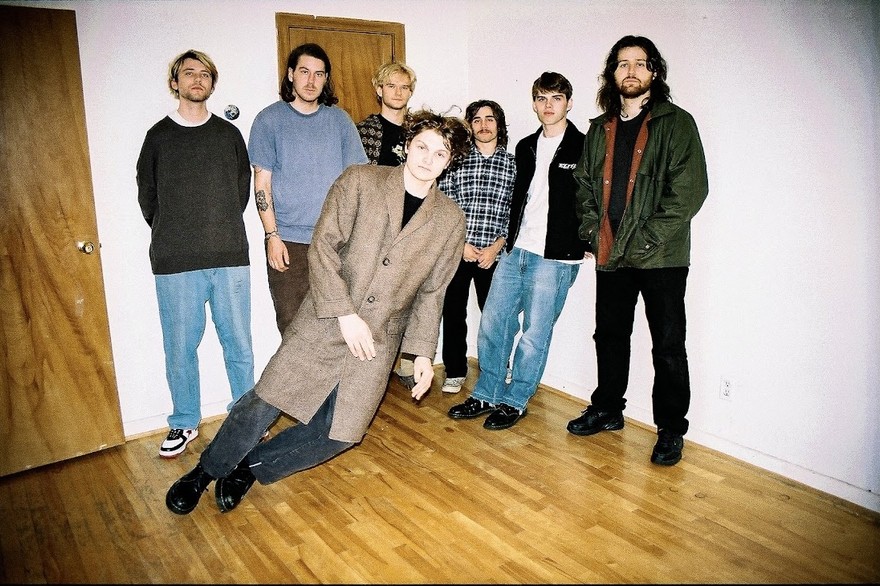 A colour photo shows seven-piece band Concrete Vehicles standing in a bedroom with hardwood floors and white walls. The member in the front is edited to look as if they are standing on a bizarre tilt as if they're falling.