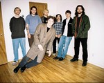 A colour photo shows seven-piece band Concrete Vehicles standing in a bedroom with hardwood floors and white walls. The member in the front is edited to look as if they are standing on a bizarre tilt as if they're falling.