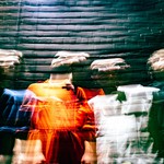 A vibrant long exposure photo shows four-piece band cute standing un front of a dark brick wall. Their faces and bodies are distorted due to the camera effect. 
