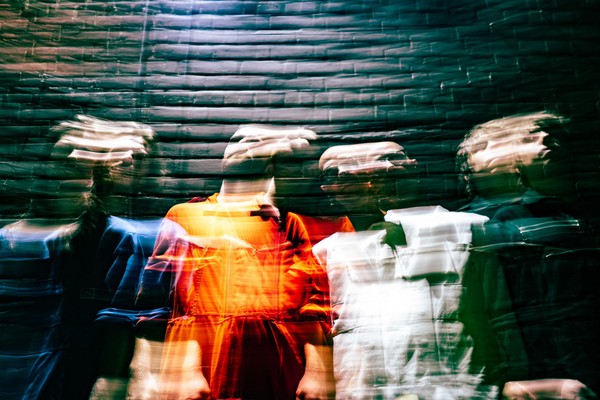 A vibrant long exposure photo shows four-piece band cute standing un front of a dark brick wall. Their faces and bodies are distorted due to the camera effect. 