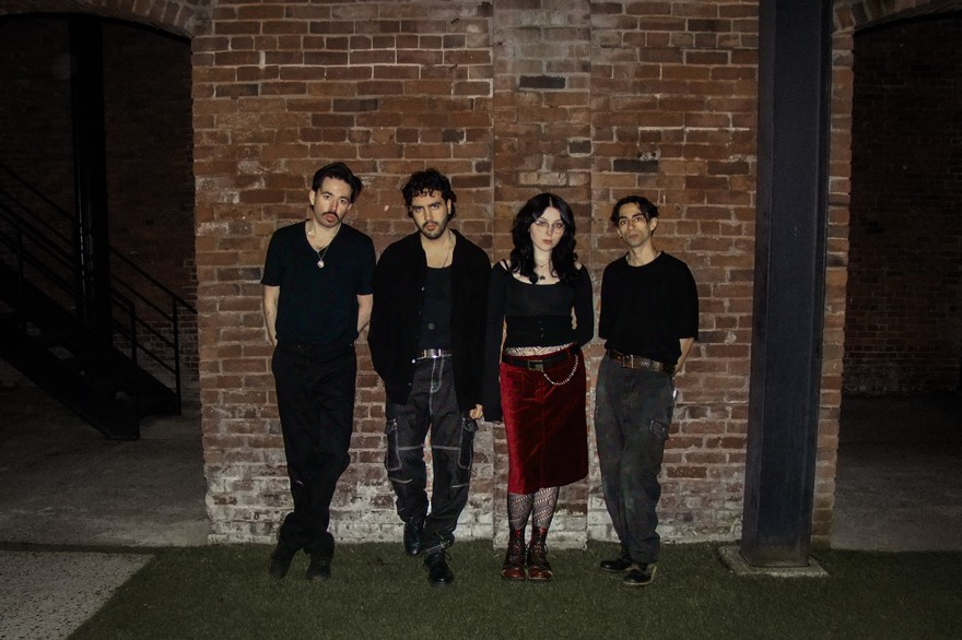 A colour photo shows four-piece group DOUR on a patch of grass standing against a brick wall at night.