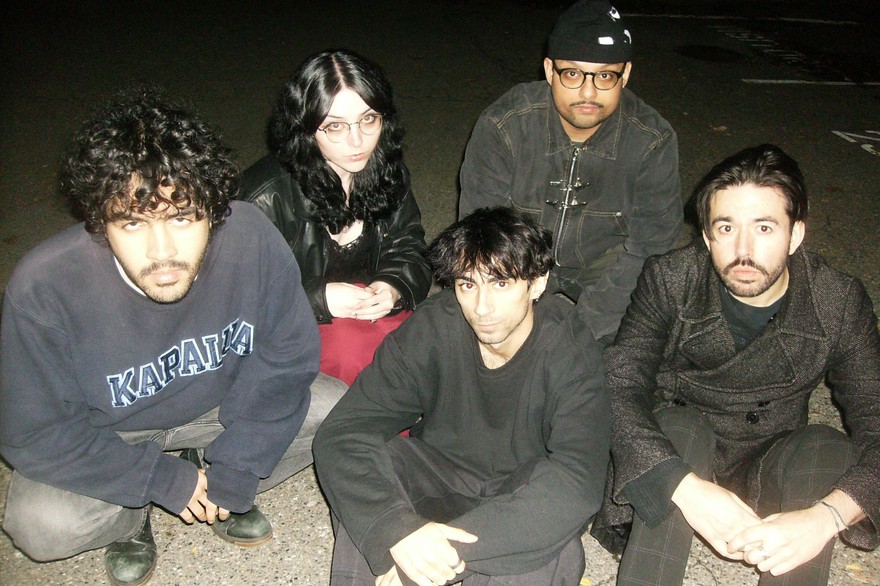 A colour photo shows five-piece group DOUR sitting in a concrete parking lots wearing dark clothes. 