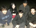 A colour photo shows five-piece group DOUR sitting in a concrete parking lots wearing dark clothes. 