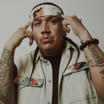 A close-up photo shows rapper Drezus standing in front of a white wall with both hands pointing to their head.