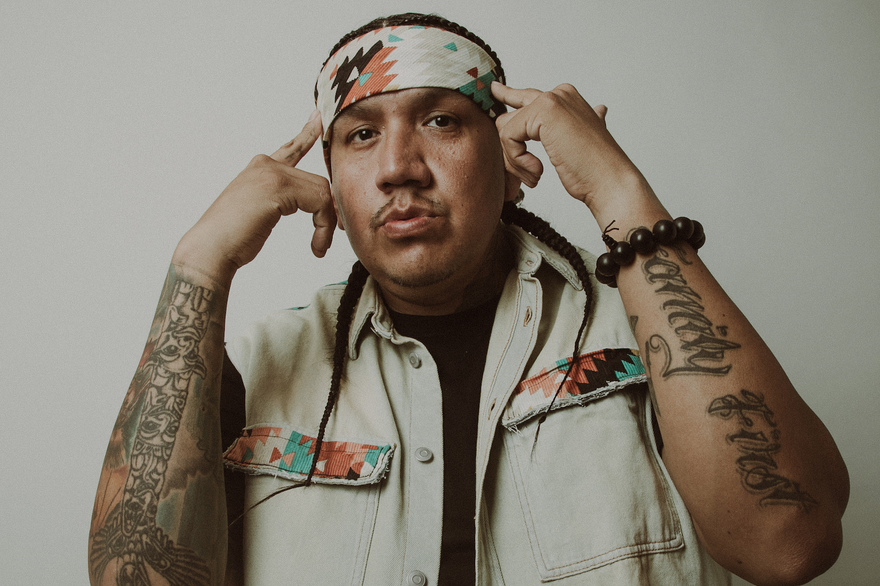 A close-up photo shows rapper Drezus standing in front of a white wall with both hands pointing to their head.