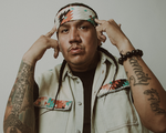 A close-up photo shows rapper Drezus standing in front of a white wall with both hands pointing to their head.