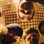 A hazy photo taken from below shows five-piece band looking down at the camera with a golden roof above that appears to be lined with egg cartons. 