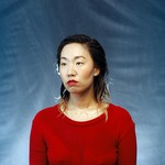 A colour photo shows artist Eejungmi posing in front of a blue backdrop wearing a bright red shirt and bright red lipstick. Their hair and face appear to be wet. 
