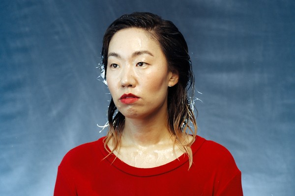 A colour photo shows artist Eejungmi posing in front of a blue backdrop wearing a bright red shirt and bright red lipstick. Their hair and face appear to be wet. 