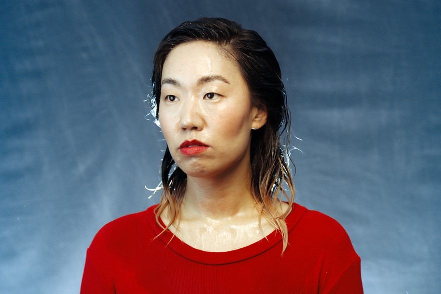 A colour photo shows artist Eejungmi posing in front of a blue backdrop wearing a bright red shirt and bright red lipstick. Their hair and face appear to be wet. 
