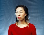A colour photo shows artist Eejungmi posing in front of a blue backdrop wearing a bright red shirt and bright red lipstick. Their hair and face appear to be wet. 