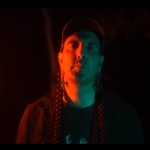 A close-up colour photo of artist ECHTHROS standing outside at night with blue and red light shining on their face. 