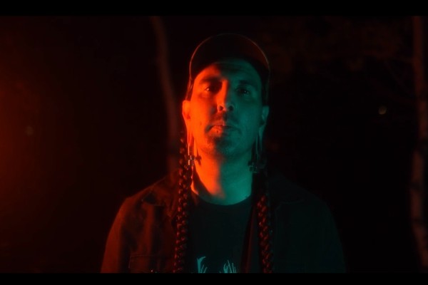 A close-up colour photo of artist ECHTHROS standing outside at night with blue and red light shining on their face. 