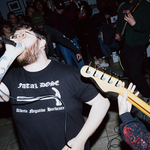 A candid colour photo shows the singer and several guitar players performing in an intimate indoor venue with the crowd standing in the background.