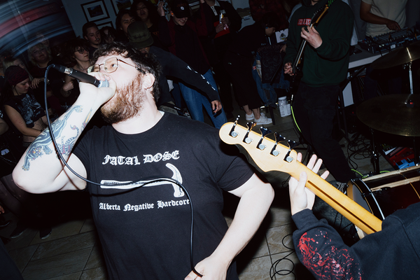 A candid colour photo shows the singer and several guitar players performing in an intimate indoor venue with the crowd standing in the background.