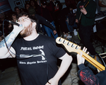 A candid colour photo shows the singer and several guitar players performing in an intimate indoor venue with the crowd standing in the background.
