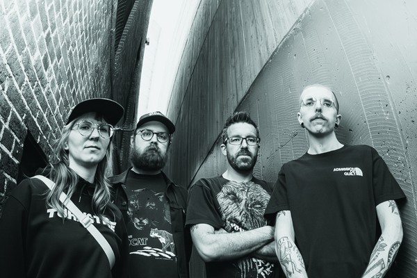 A black and white photo taken from below shows four-piece group False Body posing in an alleyway wearing black clothes. 