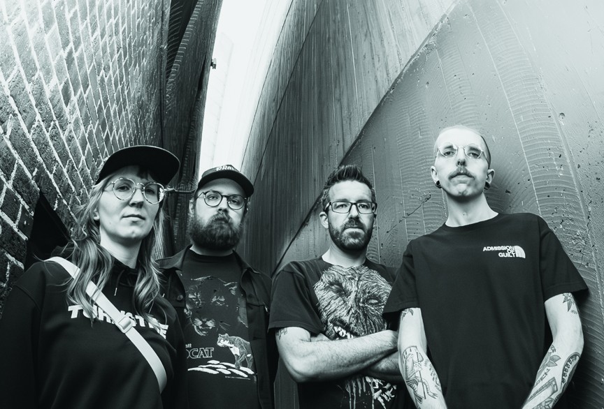 A black and white photo taken from below shows four-piece group False Body posing in an alleyway wearing black clothes. 