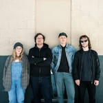 A colour photo shows four-piece group Eye of Newt posing in front of a brick wall that is white on the upper halff and painted blue on the lower half. 