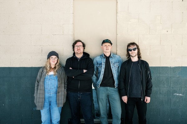 A colour photo shows four-piece group Eye of Newt posing in front of a brick wall that is white on the upper halff and painted blue on the lower half. 