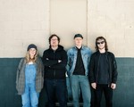 A colour photo shows four-piece group Eye of Newt posing in front of a brick wall that is white on the upper halff and painted blue on the lower half. 