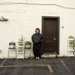 A colour photo shows the frontperson of the group Fanclubwallet standing outside of a motel room againt a white brick wall and to the left of a brown door numbered 11. Chairs and stands containing potted plants are on each side of them. 